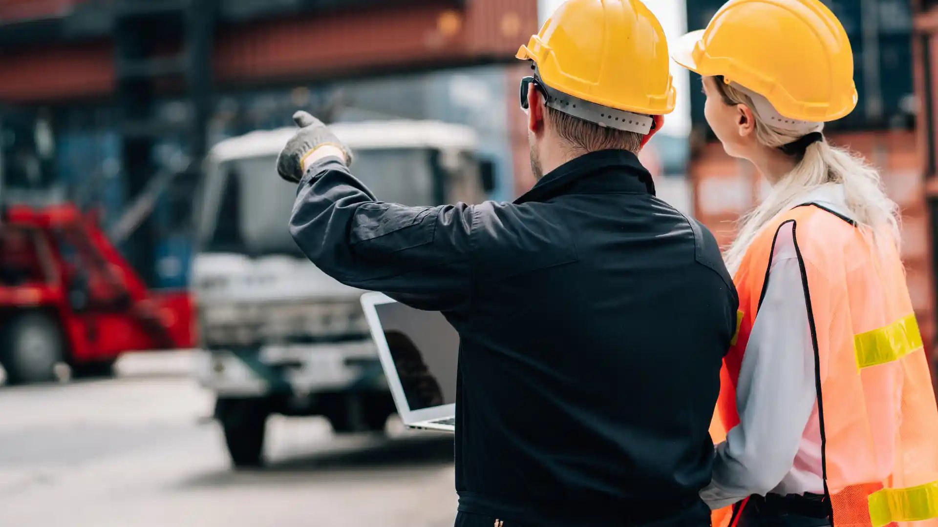 Man and woman in hard hats and hi-vis vest holding a laptop pointing to small truck in the background